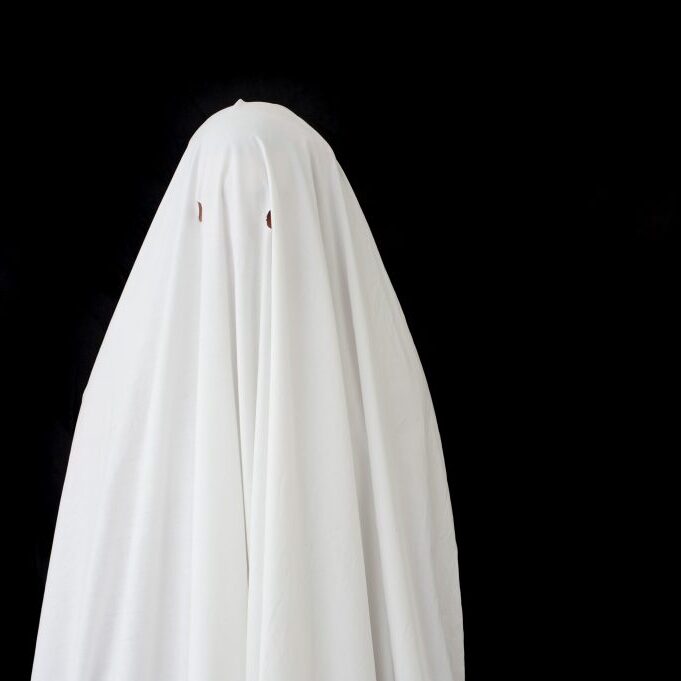 Bored person dressed up as a Halloween ghost or ghoul in a white costume standing guietly waiting against a black background